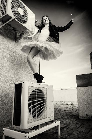 Eine Ballerina in Tutu,Jacke und Stiefeln klettert auf die Klimaanlage und posiert vor dem Himmel